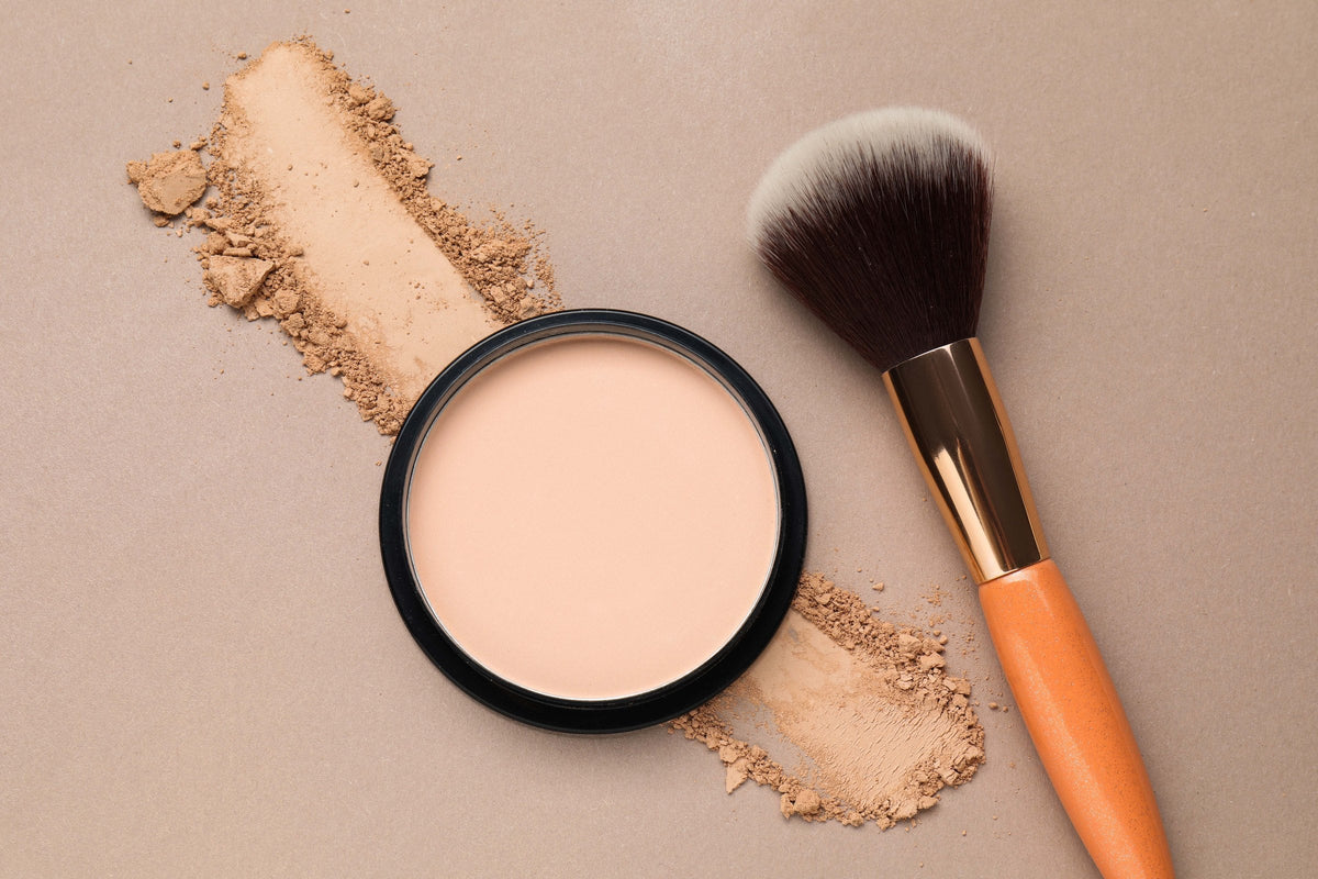 A compact of pressed face powder and a soft make-up brush on a beige background with a sweep of loose powder